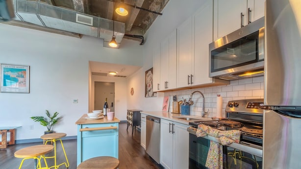 A modern kitchen with stainless steel appliances, a wooden dining area, and a bar stool. - Holiday Lofts NYC (Newark)