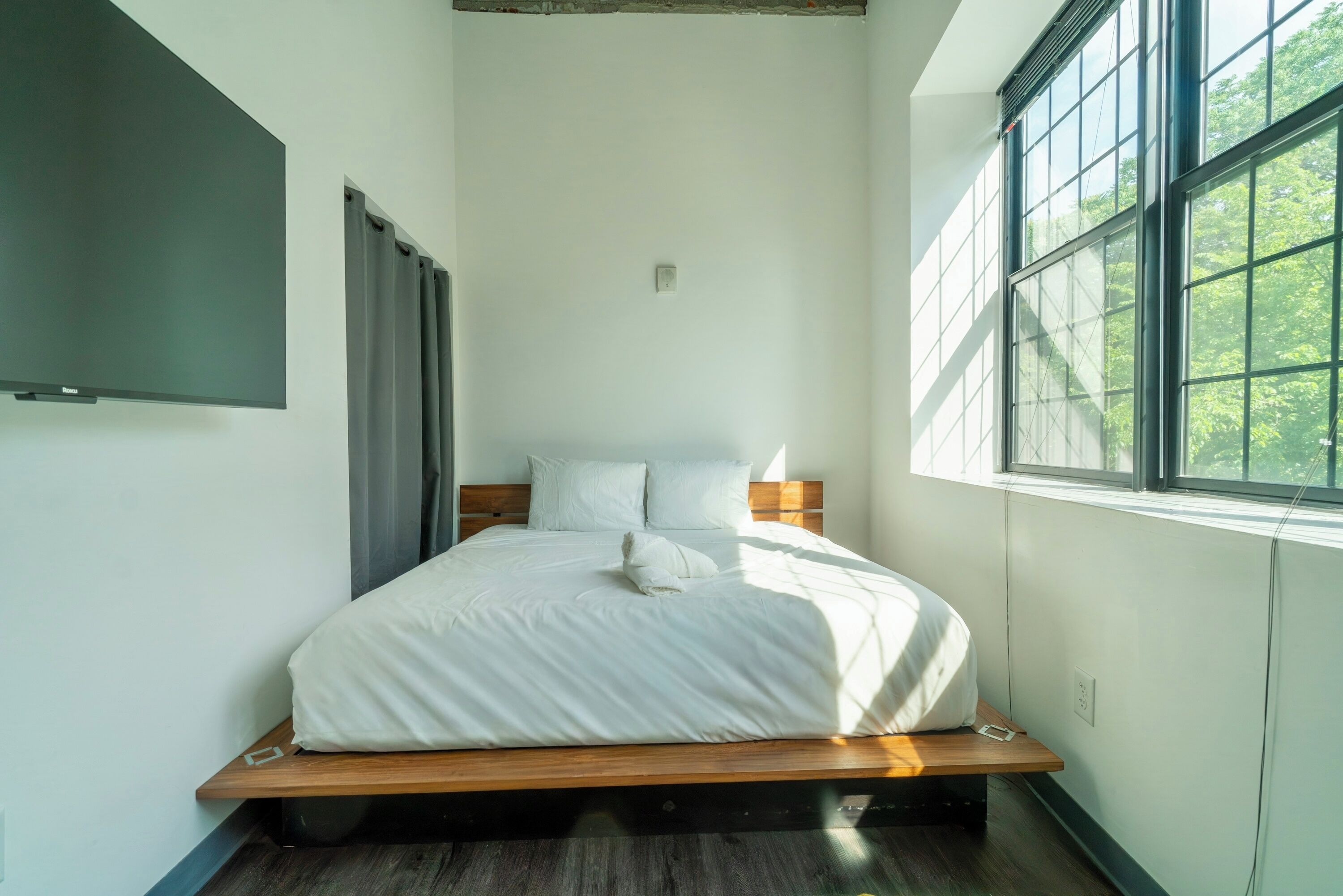 A modern bedroom with a wooden platform bed, white bedding, and a large window with a view of trees.