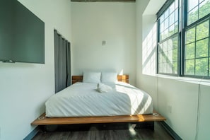 A modern bedroom with a wooden platform bed, white bedding, and a large window with a view of trees. - Holiday Lofts NYC (Newark)