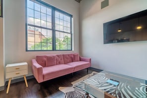 A room with a pink sofa, a small white bedside table, a glass coffee table, and a large window with a view of buildings and trees. - Holiday Lofts NYC (Newark)