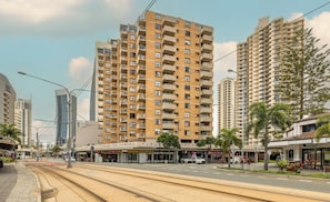Un immeuble résidentiel de grande hauteur doté de nombreux balcons, situé à proximité d’une ligne de tramway et de commerces.
