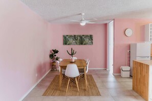 Un comedor con una mesa de madera, sillas blancas, un ventilador de techo, una planta de cactus y un reloj fijado en la pared.
