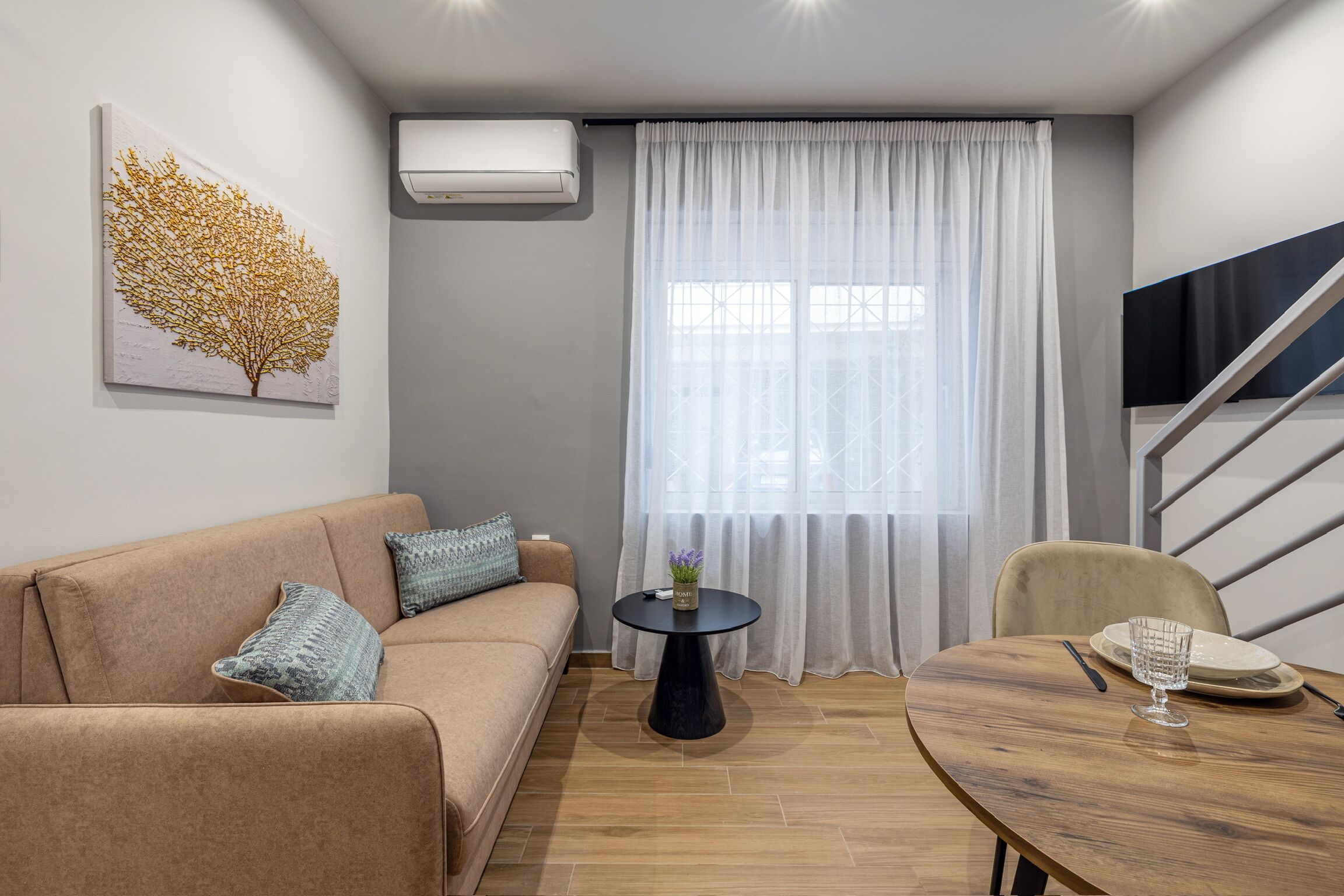 A modern living room with a sofa, a round wooden table, a small black table, and a TV mounted on the wall.