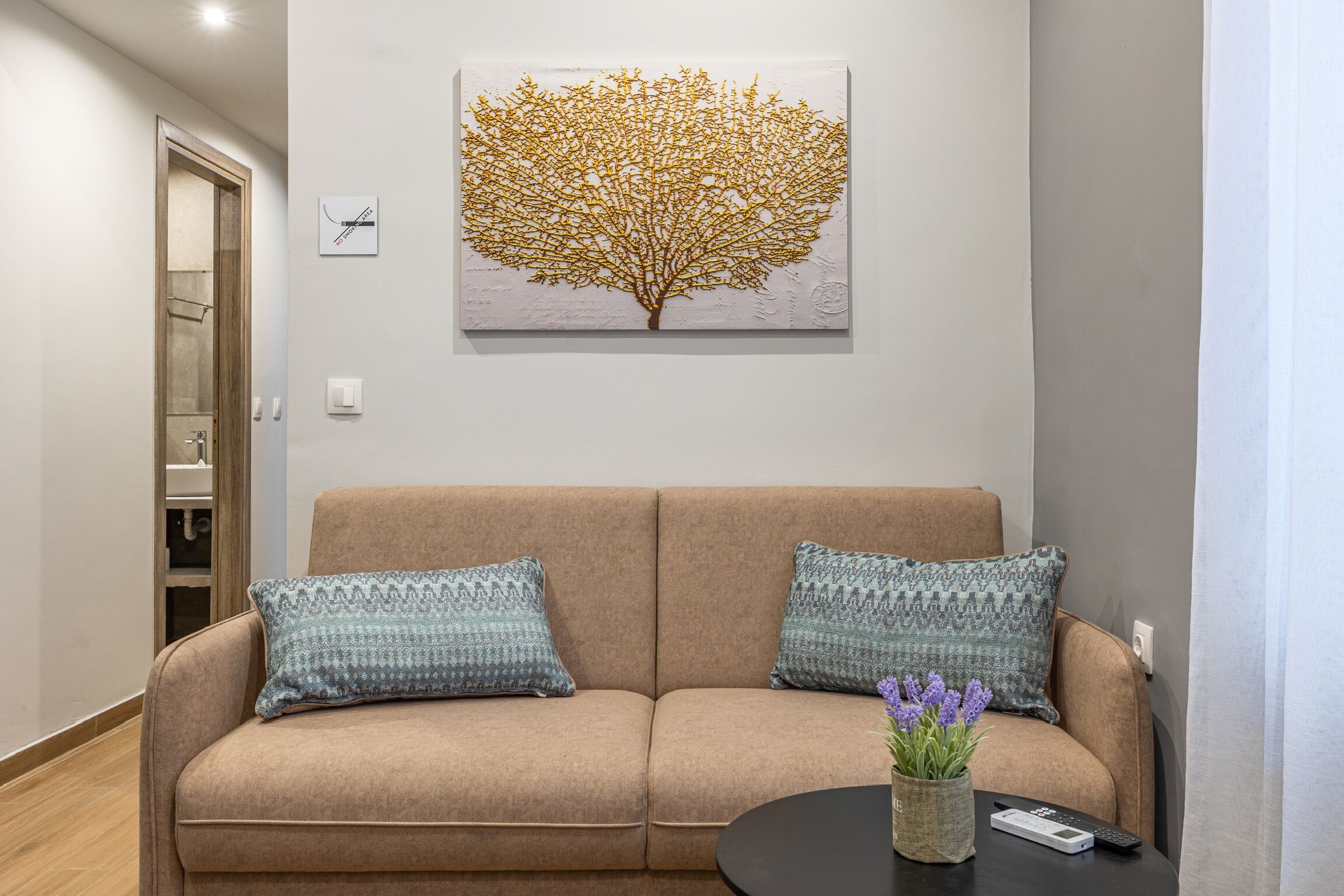Un sofá color beige con cojines estampados, un cuadro de un árbol colgado en la pared y una mesita con un control remoto y un jarrón de flores.