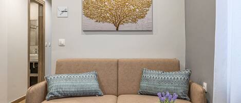 Un sofá color beige con cojines estampados, un cuadro de un árbol colgado en la pared y una mesita con un control remoto y un jarrón de flores.