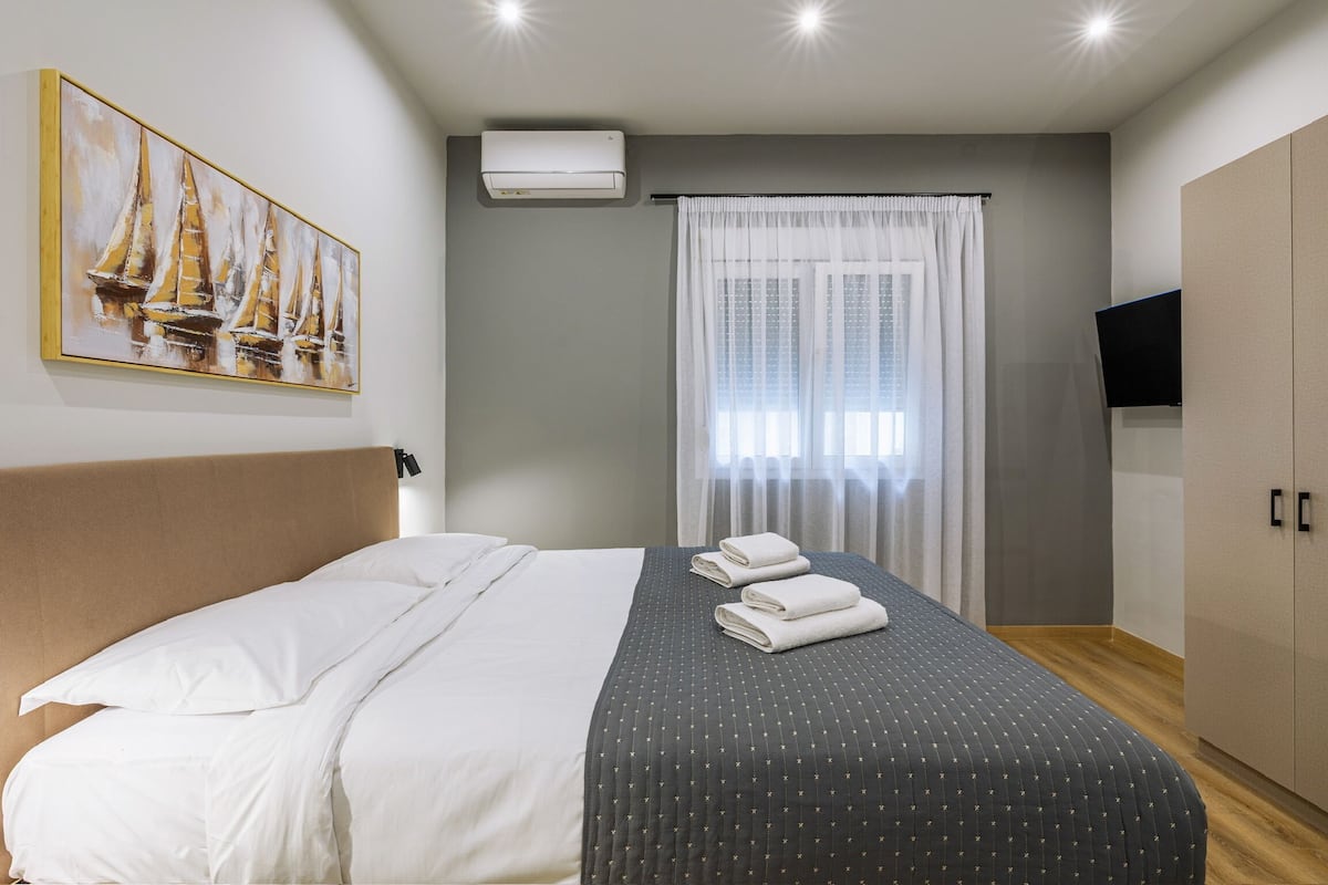 Una cama bien hecha con ropa de cama blanca y oscura, un cuadro de veleros en la pared, un televisor sobre un mueble bajo y un armario.