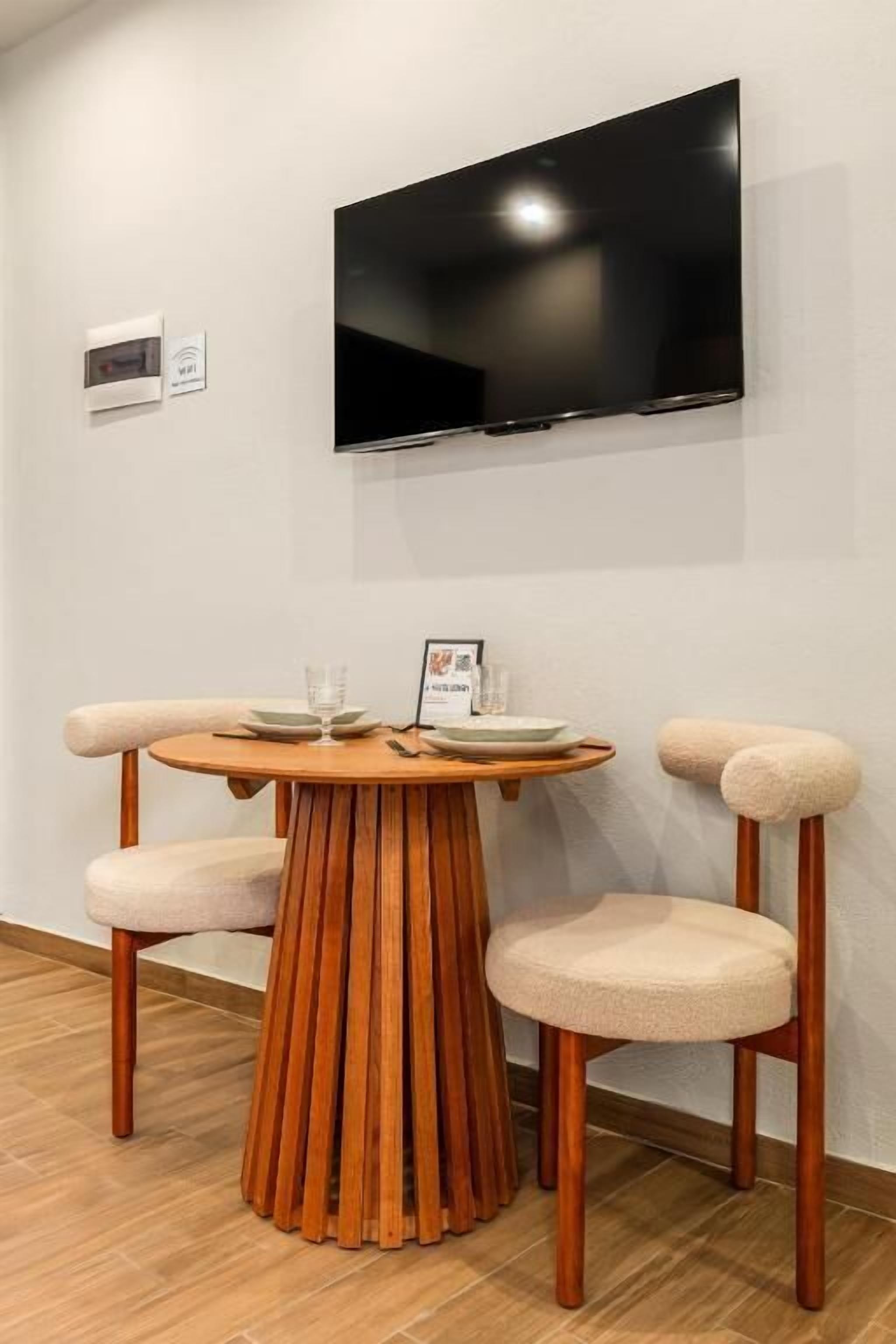A room with a round wooden table, two chairs, a flat-screen TV mounted on the wall, and a small sign on the wall.