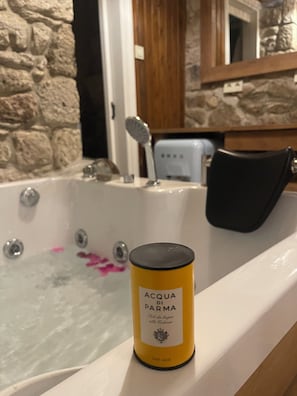 A bathtub with a bottle of ACOA DI PARMA bath salts, a wooden door, and a stone wall.