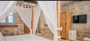 A bedroom with a stone wall, a wooden wardrobe, a TV mounted on the wall, and a bed with white bedding.