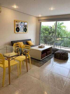 A modern living room with a glass dining table, yellow chairs, a sofa with yellow cushions, a coffee table, and a large window with a view of greenery. - Shopping del Sol Zone (Asunción)