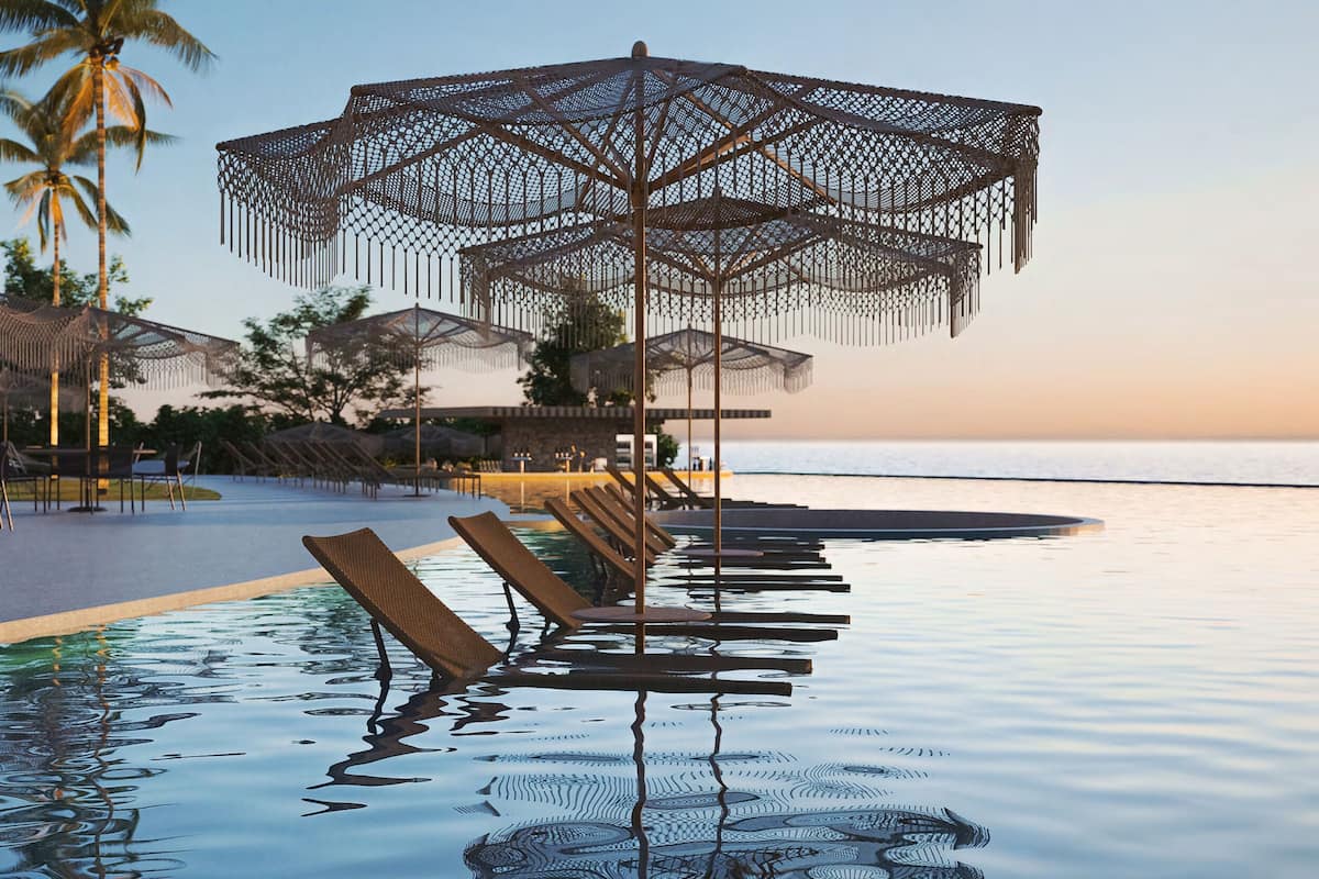 A poolside area with lounge chairs and umbrellas, palm trees, and a view of the sea.