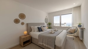 Een moderne slaapkamer met een groot bed, nachtkastjes, een stoel en uitzicht op zee.