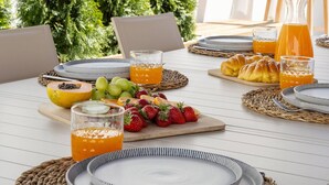 Een ontbijttafel gedekt met borden, glazen en fruit, omringd door stoelen en begroeiing.