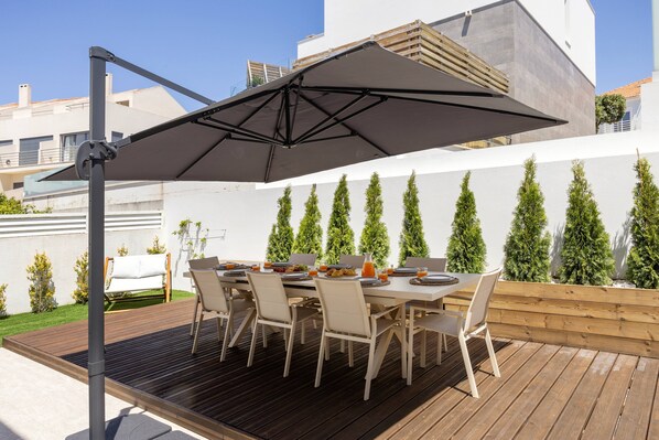 An outdoor dining area with a table set for six, surrounded by chairs, under a large umbrella. - Villa Somnium - Sunset Oasis (Lourinhã)