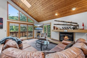 Ein geräumiges Wohnzimmer mit Ledersofas, einem Kamin, großen Fenstern und einer Holzbalkendecke.