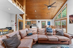 Ein geräumiges Wohnzimmer mit einem großen Ledersofa, einem Deckenventilator und Blick auf die Küche sowie den Außenbereich durch große Fenster.