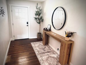 A hallway with a wooden floor, a white door, a small plant, a mirror, and a console table with a vase.