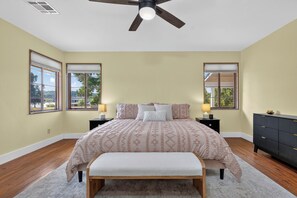 Ein Schlafzimmer mit einem Bett, zwei Nachttischen, einer Bank, einem Deckenventilator und großen Fenstern.