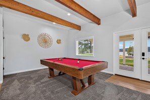Ein Billardtisch mit roter Spielvliesdecke, Holzbeinen und einer Einbuchtung in der Ecke. Dekorative Wandbilder und ein Fenster mit Jalousien sind ebenfalls zu sehen.