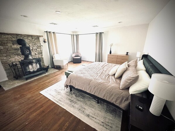 A bedroom with a stone fireplace, a bed with patterned bedding, a nightstand with a lamp, and a view of the outdoors.
