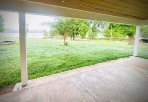 Ein überdachter Patio-Bereich mit Betonboden, umgeben von einer Rasenfläche mit Bäumen und weiter Aussicht auf ein Gewässer.