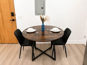 Une table de salle à manger ronde en bois, avec des chaises noires, un vase contenant des plantes séchées, ainsi que des assiettes et des couverts blancs.