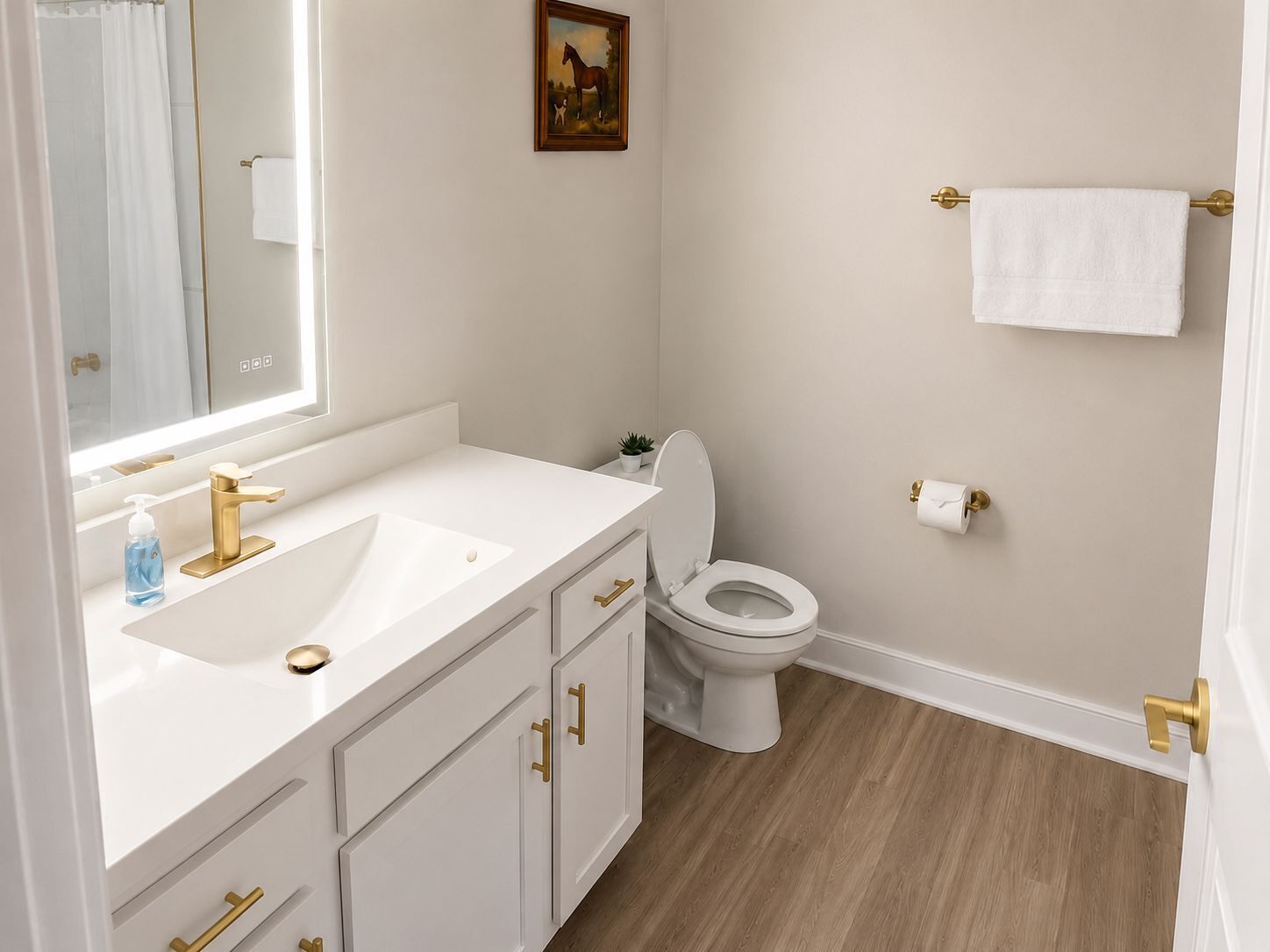 Une salle de bain avec un lavabo blanc, un robinet doré et des toilettes. On y trouve une petite plante posée sur le comptoir et un tableau accroché au mur.