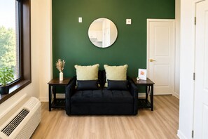 Ein Zimmer mit einer dunkelgrünen Wand, einem runden Spiegel, einer schwarzen Couch mit zwei Kissen, einem kleinen Tisch mit einer Vase und einer Pflanze sowie einer weißen Tür.