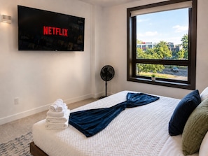 Ein Hotelzimmer mit Bett, einem an der Wand befestigten Fernseher mit dem Netflix-Logo, einem Fenster mit Blick auf einen Parkplatz und Bäume sowie einem Standventilator.