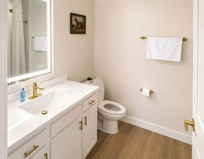 Ein Badezimmer mit einem weißen Waschbecken, einem goldenen Wasserhahn und einer Toilette. An der Wand hängt ein gerahmtes Bild und es gibt einen Handtuchhalter.