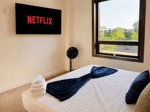 A bedroom with a bed, a wall-mounted TV displaying 'NETFLIX', a window with a view of a parking lot, and a standing fan. - Downtown! Walk to 4th Street Live, Brown Theater (Louisville)