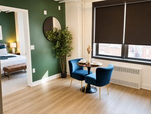 Ein Zimmer mit einem Bett, einem runden Tisch mit Stühlen, einer Topfpflanze und einem Fenster mit Jalousien.