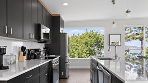 Une cuisine moderne avec des meubles de couleur sombre, des appareils électroménagers en acier inoxydable et une grande fenêtre donnant sur des arbres et un plan d’eau.