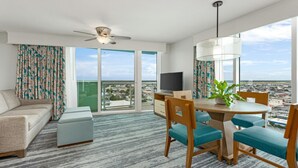 Ein modernes Hotelzimmer mit Balkon, einem Essbereich mit Tisch und Stühlen, einer Couch, einem Fernseher und einem Deckenventilator.