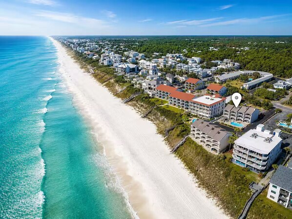 An aerial view of a coastal area with a beach, residential buildings, and a hotel with multiple floors and balconies.