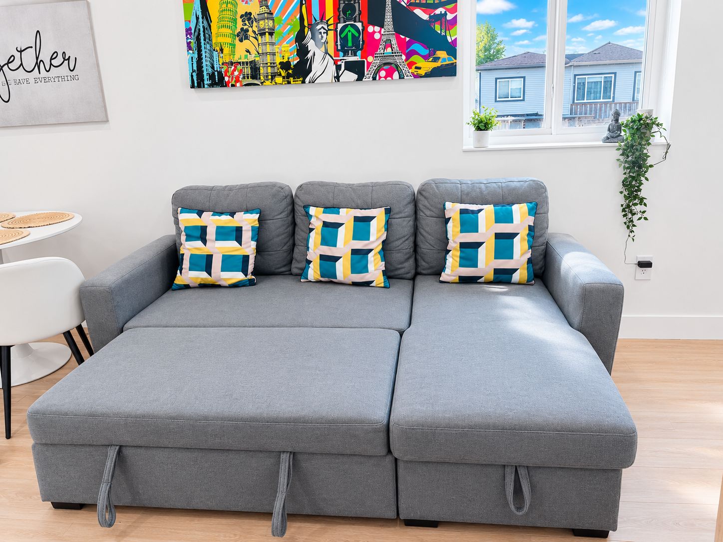 Ein graues Ecksofa mit geometrischen Kissen, ein weißer runder Tisch mit gewebten Platzsets und ein großes abstraktes Gemälde über der Couch.