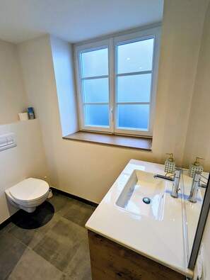 Une salle de bain comprenant des toilettes, un lavabo avec deux robinets et une fenêtre à carreaux au-dessus du lavabo.