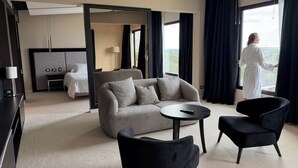 Una habitación de hotel moderna con sofá, dos sillones, una mesita redonda, cama, televisor y un ventanal con cortinas.