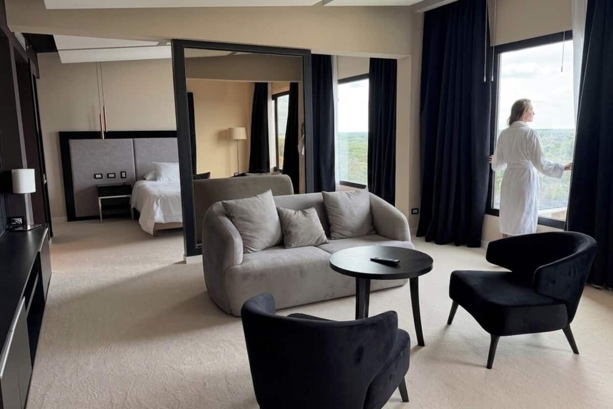 Una habitación de hotel moderna con sofá, dos sillones, una mesita redonda, cama, televisor y un ventanal con cortinas.