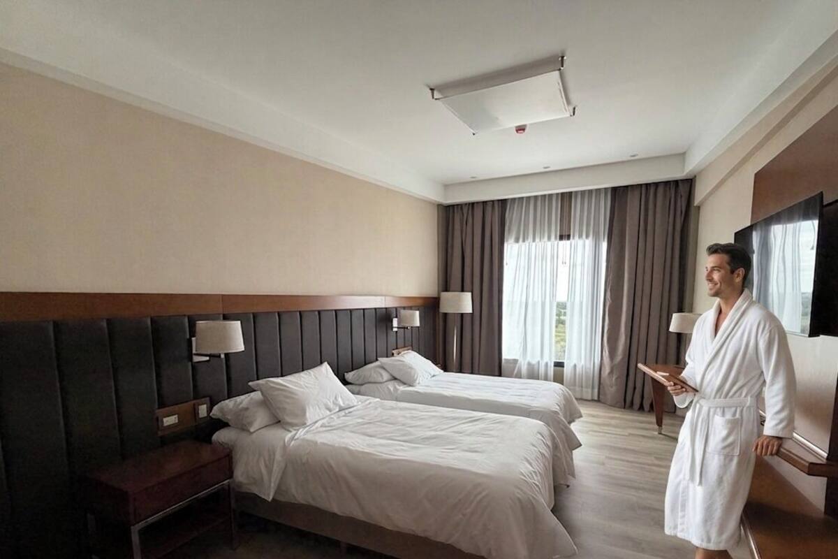 Habitación de hotel con dos camas, un escritorio, una silla, un banco y un ventanal con cortinas.