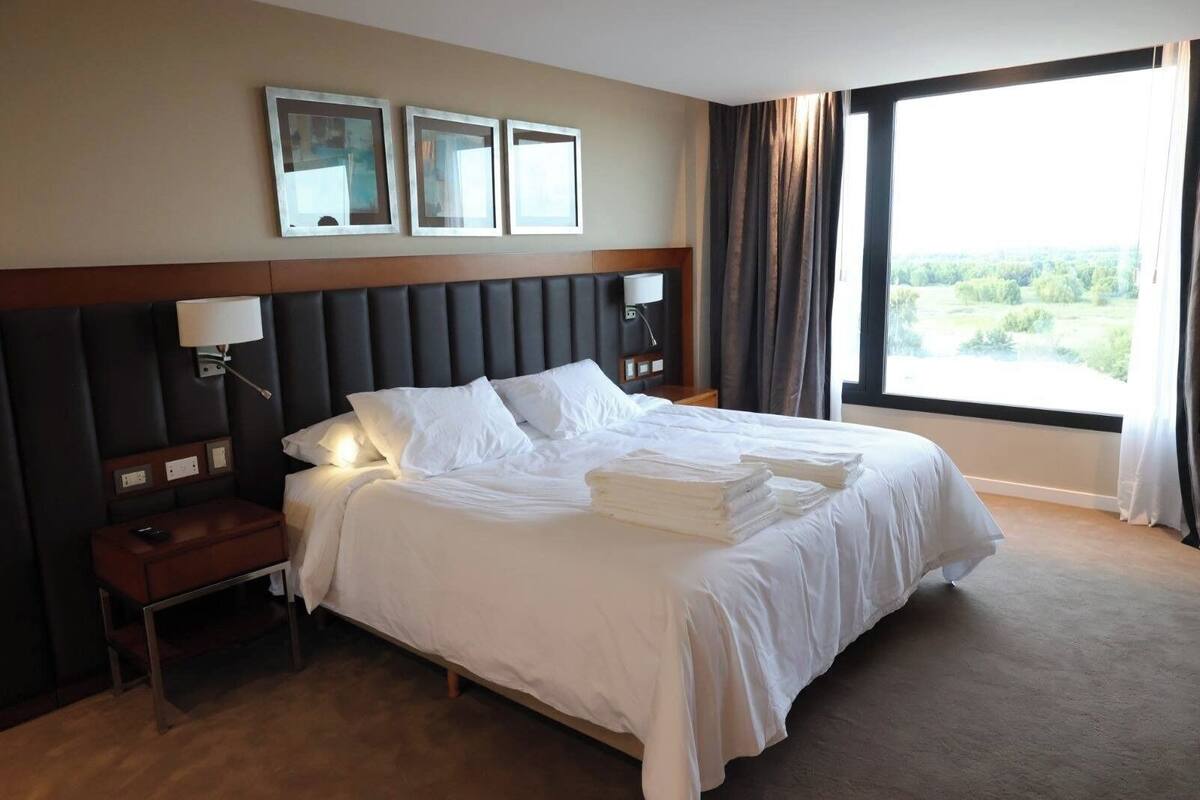 Una habitación de hotel con una cama grande, una mesita de noche, una lámpara y un ventanal con cortinas.