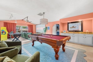 A pool table in a room with a sofa, a foosball table, and a TV screen displaying 'MOVIE NIGHT'. - Pool, Hot Tub, PickleBall, Firepit, 5 min to IMG (Bradenton)