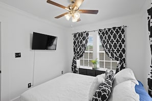 Ein Schlafzimmer mit einem Bett, einem Deckenventilator, einem an der Wand montierten Fernseher und gemusterten Vorhängen.