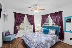 A bedroom with a bed, a chair, a ceiling fan, and a window with curtains. - Magnolia Beach Cottage (Gulfport)