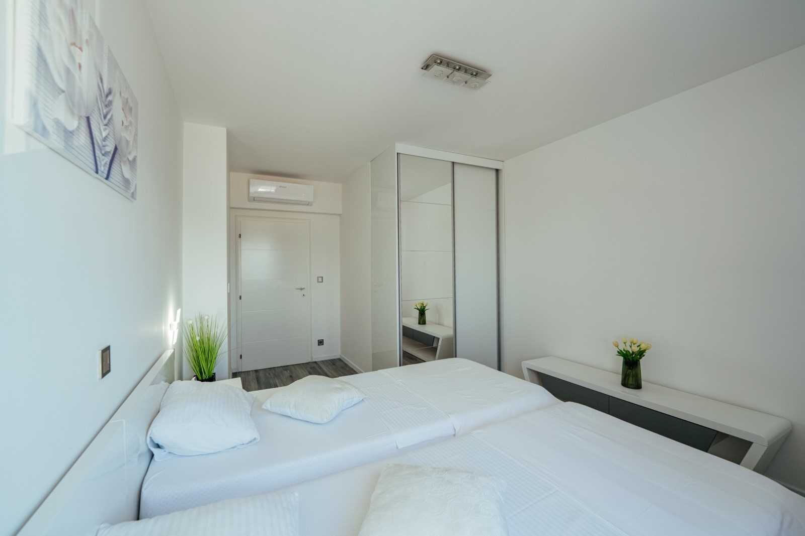 A modern hotel room with a bed, bedside tables, a mirror, a small plant, and a door leading to another room.