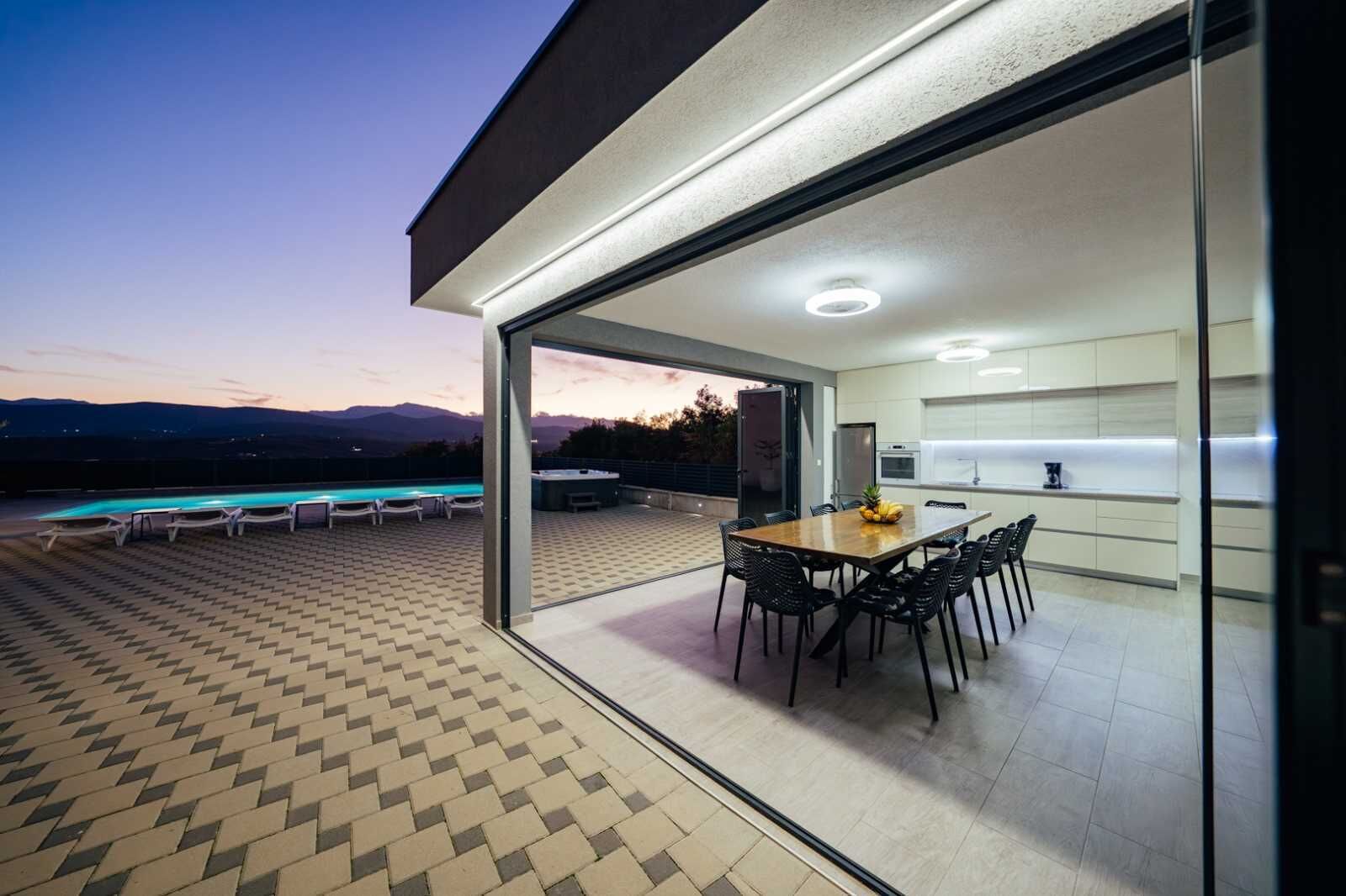 A modern outdoor dining area with a pool, a kitchen with white cabinets, and a view of mountains.