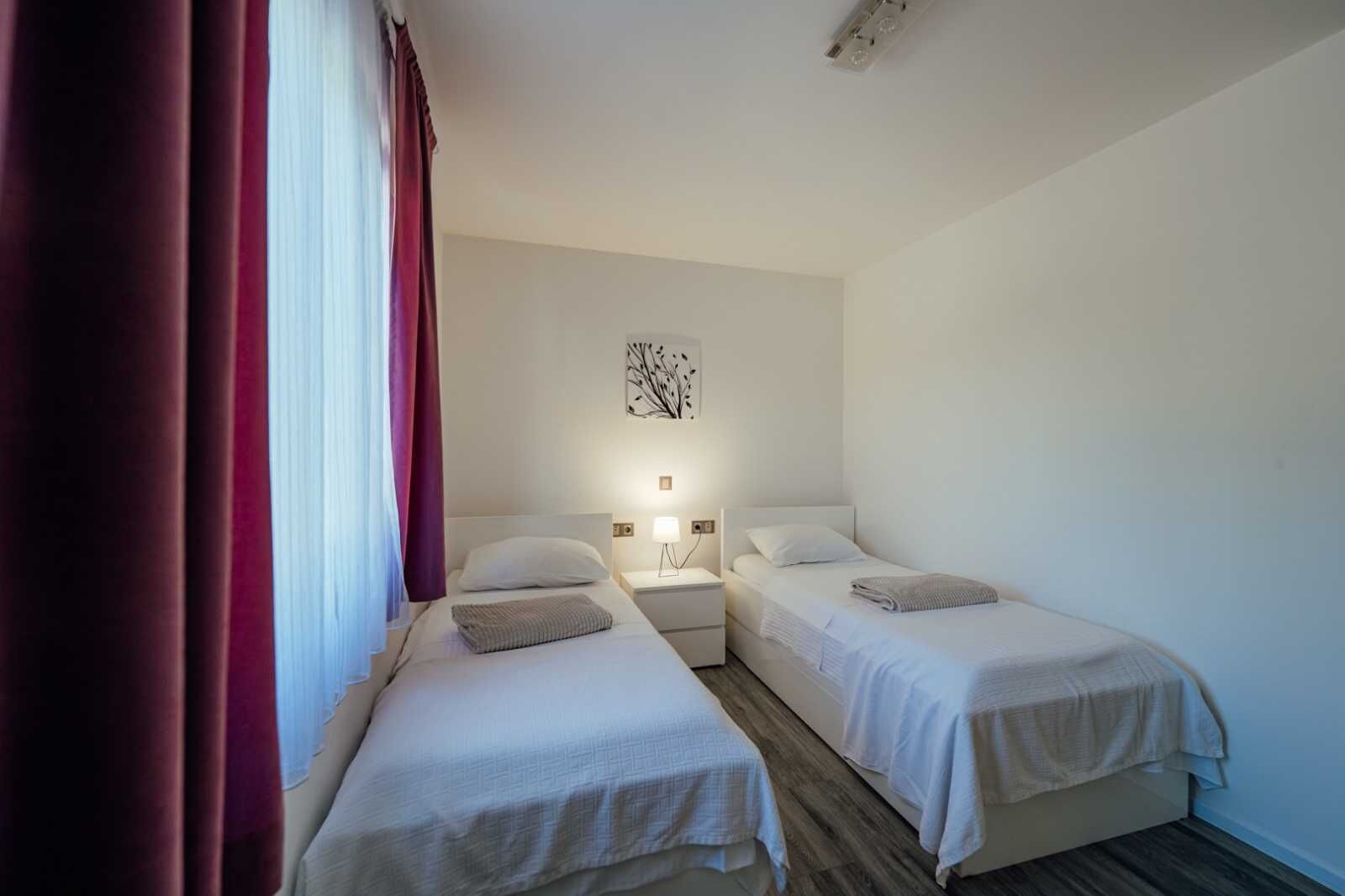 A hotel room with two single beds, a nightstand with a lamp, a wall art piece, and a window with curtains.