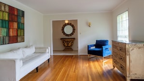 Ein Zimmer mit einer weißen Couch, einem blauen Sessel, einem Holzkabinett, einem Spiegel und einem farbenfrohen abstrakten Gemälde.