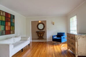 Ein Zimmer mit einer weißen Couch, einem blauen Sessel, einem Holzkabinett, einem Spiegel und einem farbenfrohen abstrakten Gemälde.