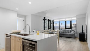 A modern kitchen with a built-in dishwasher, a large island, and a dining area with a sofa and a TV stand. - Premiere Lakeshore Living with excellent amenities. Dog friendly (Kelowna)
